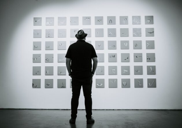 Scott Bennett standing in front of a wall-mounted grid of abstract square artworks. A calm, monochrome scene exploring repetition, form, and minimalist composition.