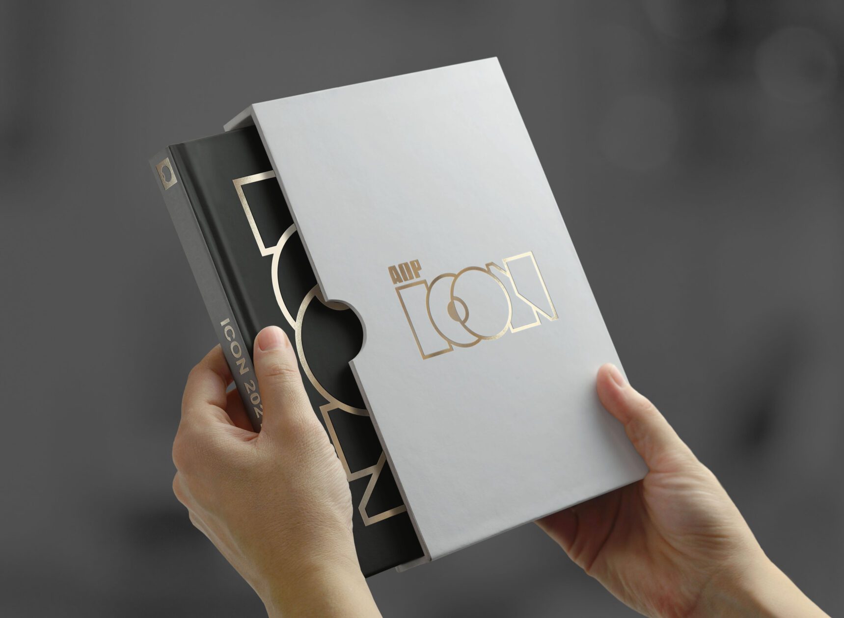 Hands holding the AOP ICON 2021 award book, partially removed from a white slipcase with gold foil logo detailing.